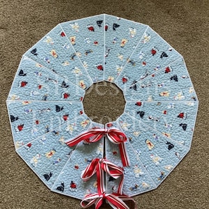 May include: A light blue quilted Christmas tree skirt with a repeating pattern of small figures and bows. The skirt has a circular shape with a center opening and is decorated with red and white striped bows.