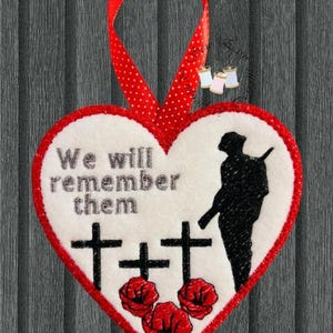 May include: Heart-shaped decoration with a red border and a red polka-dot ribbon. The ornament features the text "We will remember them", three black crosses, a soldier silhouette, and three red poppies.