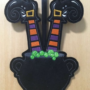 May include: A Halloween-themed felt decoration featuring a black cauldron with witch legs and feet. The legs are striped orange and purple, with black boots and gold buckles. Green bubbles spill from the cauldron. The text "SB Designs embroidery" is visible.