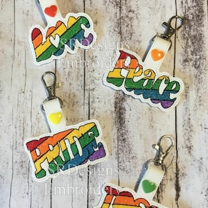 May include: Four rainbow-colored keychains with the words "Love", "Peace", "Pride", and "I Love You" in a retro font. Each keychain has a small heart charm in a different color.