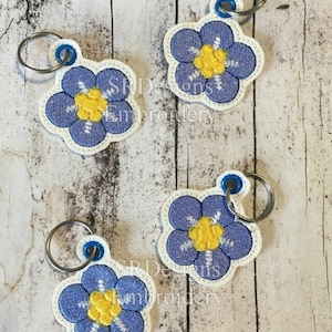 May include: Four blue and white forget-me-not flower keychains with yellow centers. Each keychain has a silver ring.