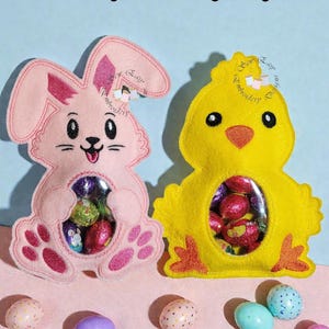 Può includere: Coniglietto in feltro rosa e pulcino in feltro giallo porta caramelle pasquali pieni di ovetti di cioccolato. Il coniglietto e il pulcino hanno finestre di plastica trasparente. L'immagine mostra anche uova di Pasqua decorative. Il testo recita "Sew easy embroidery designs."