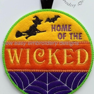 May include: A round felt hanging ornament with a black ribbon loop. The top section is yellow with a black silhouette of a witch on a broom and a bat. The text reads HOME OF THE WICKED. The middle is orange, and the bottom is purple with a white spiderweb.