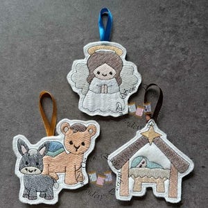 May include: Embroidered Christmas ornaments featuring an angel, a camel with a donkey, and a manger scene with baby Jesus. Each ornament has a ribbon for hanging. The angel has a halo and is wearing a white dress. The camel and donkey are tan and gray.