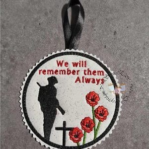 May include: A round, white ornament with a black scalloped edge. It features a black silhouette of a soldier, a cross, and three red poppies. The text "We will remember them Always" is in red. A black ribbon is attached for hanging.