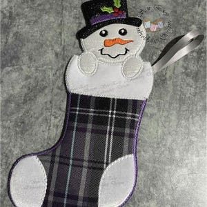 May include: Embroidered Christmas stocking featuring a snowman design. The snowman has a black top hat with holly, an orange carrot nose, and a smiling face. The stocking has a plaid pattern in shades of purple, gray, and white, with a white cuff and toe.