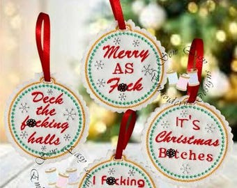Op de afbeelding: Vier witte kerstversieringen met rode linten. Elke cirkelvormige versiering heeft een geschulpte rand, groene en gouden stiksels en rode tekst. De tekst op de versieringen luidt: "Merry As Fuck", "Deck the focking halls", "It's Christmas Bitches" en "I focking HATE Christmas".