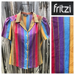 May include: A striped button-up shirt with a collar. The shirt has vertical stripes in shades of red, orange, yellow, blue, purple, and brown. The shirt is labelled "fritzi" on the collar.