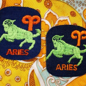 Op de afbeelding: Twee geborduurde Aries-patches. Elke patch heeft een groene ram, een oranje Aries-symbool en het woord "ARIES" in oranje draad op een marineblauwe achtergrond. De patches zijn geplaatst op een kleurrijke stof met patronen.