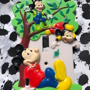 May include: A light switch cover featuring three Mickey Mouse characters. The cover is made of plastic and has a white background. The Mickey Mouse characters are in various poses, including one sitting on a green branch, one climbing a tree, and one sitting on the ground. The cover is decorated with green leaves and a brown tree trunk.