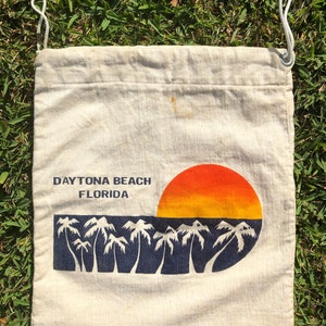 May include: A white canvas drawstring bag with the text "DAYTONA BEACH FLORIDA" and a graphic of a sunset over palm trees.