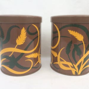 May include: Two brown metal canisters with a yellow and green floral design. The canisters have a brown lid.
