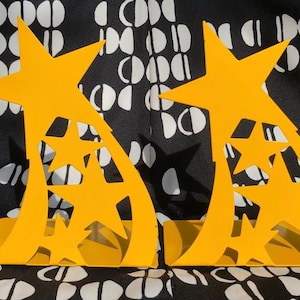 May include: A pair of yellow metal bookends shaped like shooting stars with cut-out stars. The bookends are on a black and white patterned surface.