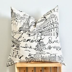 May include: Cushion with a black and white Parisian cityscape design. The cushion features iconic landmarks like the Eiffel Tower and Arc de Triomphe, with the words "L'amour Paris" in script.