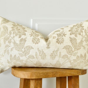May include: A rectangular cushion with a floral pattern in beige and tan hues. The cushion is placed on a small, round wooden stool. The floral design features stylized flowers and leaves.