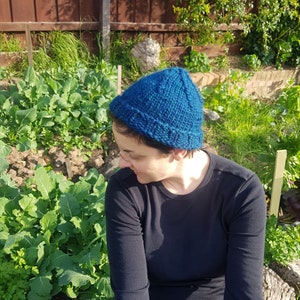 May include: A person wearing a blue knitted beanie hat, sitting in a garden setting. The hat has a textured pattern and is pulled down over the person's ears.