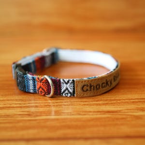 May include: A colorful dog collar with a patterned fabric in shades of blue, orange, and black. The collar has a brown leather patch with the text "Chocky Rib" and a silver metal ring.