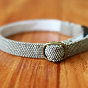 May include: A beige fabric cat collar with a brass buckle. The collar is adjustable and has a safety release buckle.