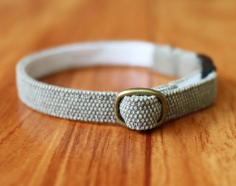 May include: A beige fabric cat collar with a brass buckle. The collar is adjustable and has a safety release buckle.