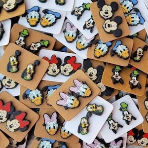 May include: A collection of colorful character shoe charms featuring Mickey Mouse, Minnie Mouse, Donald Duck, Daisy Duck, and Goofy. The charms are attached to small brown and white cards, showcasing the iconic Disney characters in various poses.