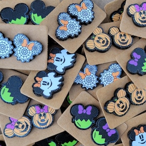 May include: A collection of mouse-shaped stud earrings on brown cardboard display cards. Designs include orange pumpkins, white ghosts, green Frankenstein-style faces, and spider webs, all featuring black glitter accents and small colorful bows.