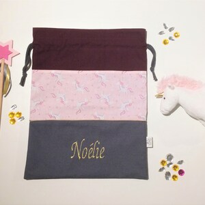 May include: A grey drawstring bag with a pink unicorn print and a burgundy top. The bag has the name "Noélie" embroidered on the front in gold thread.