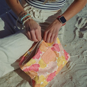 May include: A floral print cosmetic bag with a zipper closure. The bag features a pattern of pink, orange, and yellow hibiscus flowers. A leather patch with the text "Sunshine Luxe" is sewn on the bag.