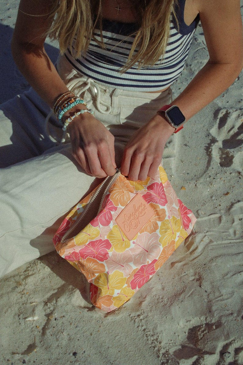 May include: A floral print cosmetic bag with a zipper closure. The bag features a pattern of pink, orange, and yellow hibiscus flowers. A leather patch with the text "Sunshine Luxe" is sewn on the bag.