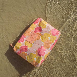 May include: A rectangular floral print pouch with a pink, yellow, and orange hibiscus pattern. The pouch has a small, rectangular label with the text "Simply Sophie". The pouch is resting on a sandy surface with water.