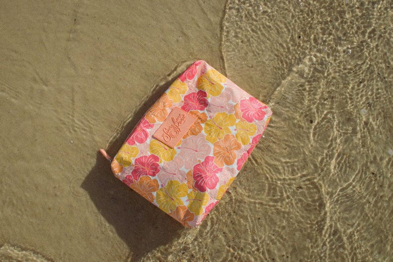 May include: A rectangular floral print pouch with a pink, yellow, and orange hibiscus pattern. The pouch has a small, rectangular label with the text "Simply Sophie". The pouch is resting on a sandy surface with water.