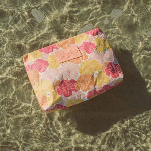 May include: A rectangular cosmetic bag with a floral pattern of pink, yellow, and orange hibiscus flowers. The bag has a light pink label with the text "Sunshine State". The bag is floating in clear water.