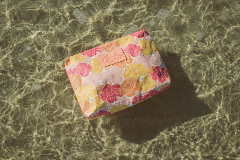 May include: A rectangular cosmetic bag with a floral pattern of pink, yellow, and orange hibiscus flowers. The bag has a light pink label with the text "Sunshine State". The bag is floating in clear water.