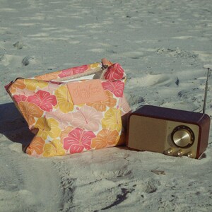 May include: A floral print bag and a retro-style radio sit on a sandy surface. The bag features a pink, orange, and yellow hibiscus pattern. The radio has a brown wooden exterior and a silver dial.