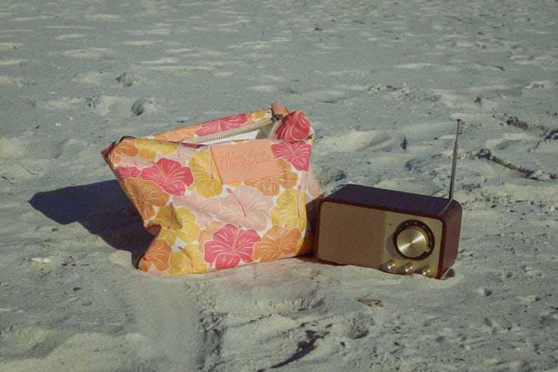 May include: A floral print bag and a retro-style radio sit on a sandy surface. The bag features a pink, orange, and yellow hibiscus pattern. The radio has a brown wooden exterior and a silver dial.