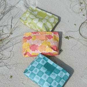 May include: Three colorful cosmetic bags with tropical patterns rest on sand. The bags feature a green leaf print, a floral pattern in pink, yellow, and orange, and a blue and white checkered design. Each bag has a leather label.