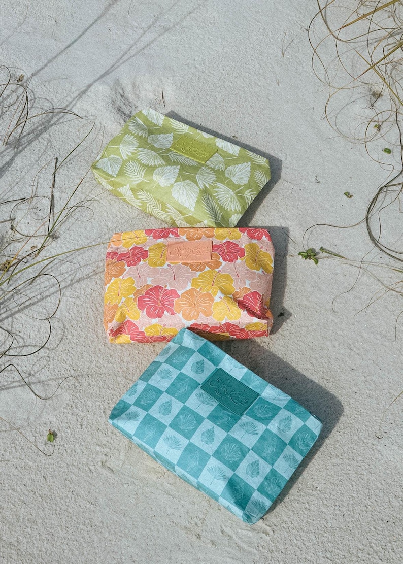 May include: Three colorful cosmetic bags with tropical patterns rest on sand. The bags feature a green leaf print, a floral pattern in pink, yellow, and orange, and a blue and white checkered design. Each bag has a leather label.