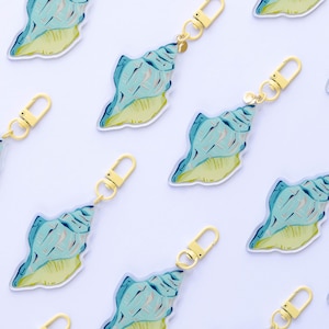 May include: Keychains featuring a seashell design. Each keychain has a gold-colored clasp and a seashell charm with blue and yellow hues. The seashells are arranged on a white background.