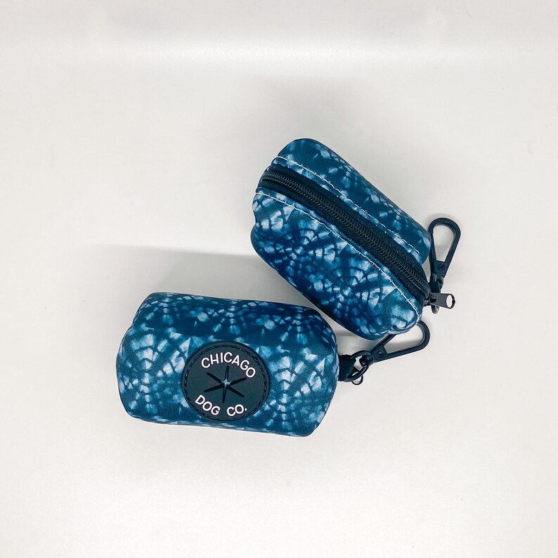 May include: Two blue and white tie-dye dog poop bag holders with a black zipper and a black carabiner clip. The holder on the left has a black circle with the text "CHICAGO DOG CO."