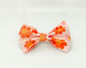 May include: A fabric bow tie featuring a light pink background with a repeating pattern of orange and yellow daisy-like flowers. The bow is constructed with a central fabric knot and pleated sides, creating a structured, symmetrical shape.