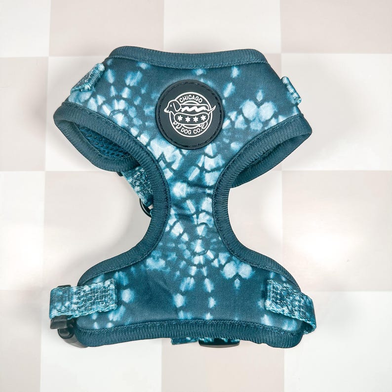 May include: A teal blue dog harness with a tie-dye pattern. The harness has a black buckle and a circular patch with the text "CHICAGO  PY DOG CO."