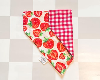 May include: A triangular pet bandana featuring a cream background with a red strawberry print on one side and a red and white checkered pattern on the other. A small white fabric tag with black text is attached to the bottom edge of the bandana.