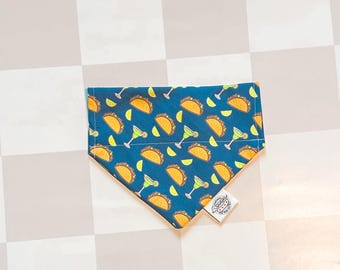 May include: A triangular pet bandana in a deep blue fabric featuring a repeating pattern of yellow tacos, green margarita glasses, and lime wedges. A small white fabric tag with a black graphic logo is attached to the bottom right corner.