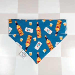 May include: Blue dog bandana featuring a pattern of amber beer bottles, white beer cans, and yellow shot glasses. The bandana has a white tag with black text. The design is repeated across the fabric, creating a fun, themed accessory.