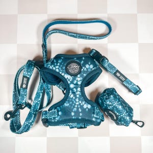 May include: A blue and white tie-dye dog harness, lead, and poop bag holder set. The harness has a black buckle and a white logo that says "Chicago Dog Co.". The lead is blue and white tie-dye and has a black clip. The poop bag holder is blue and white tie-dye and has a black clip.