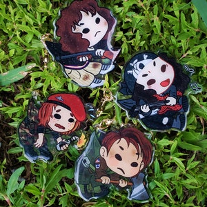 May include: Four colorful cartoon character keychains with different expressions. Each keychain features a character with a different weapon, including a rifle, a guitar, a grenade, and an axe. The keychains are made of clear plastic with glitter accents.