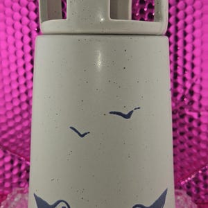 May include: A ceramic candle holder with a light beige speckled finish. The cylindrical base features a blue bird and wave design. The top section has a square cut-out design. The candle holder is approximately 8 inches tall.