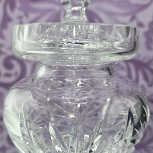 May include: A clear, cut-glass jar with a lid. The jar has a rounded body and a faceted lid with a knob. The glass is intricately cut with a starburst pattern on the base and vertical cuts on the sides. The jar is designed to hold small items.