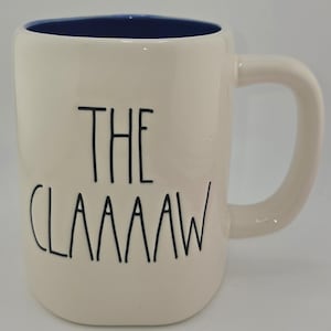May include: Cream-colored ceramic mug with a blue interior and a curved handle. The mug features the text "THE CLAAAAAW" in black, stylized lettering. A simple, everyday coffee or tea mug.