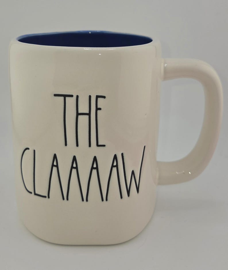 May include: Cream-colored ceramic mug with a blue interior and a curved handle. The mug features the text "THE CLAAAAAW" in black, stylized lettering. A simple, everyday coffee or tea mug.