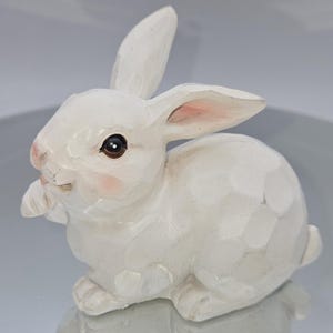 May include: A white bunny figurine with a carved wood appearance. The bunny has pink accents on its ears and cheeks, and dark brown eyes. The bunny is sitting with its paws together, and its ears are perked up. The figurine is a decorative item.